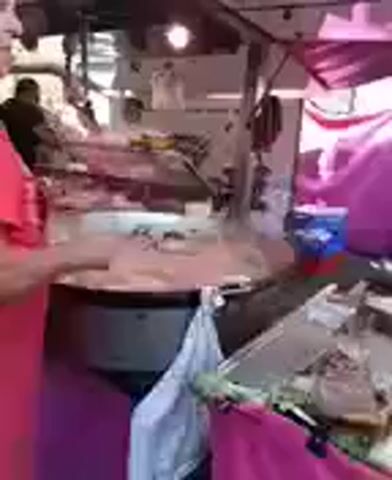 A person in a red shirt is at a market stall. They are holding a circular object, possibly food, over a large round pan. The stall is covered by a canopy, and there are other people and market goods visible in the background.