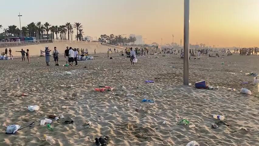 This media file captures a beach scene in Barcelona, Spain, at sunset. The beach is heavily littered with various types of trash, including plastic bottles, cups, food wrappers, and bags, strewn across the sand. Numerous people are present on the beach, some sitting or lying down, others walking or standing in groups. Many appear to be enjoying the end of the day, with the warm glow of the setting sun casting long shadows. The atmosphere seems relaxed despite the significant amount of litter. The presence of palm trees and urban buildings in the background provides clear location cues for Barcelona. The sky is clear with a soft, orange hue from the setting sun, indicating fair weather. No visible text is present in the media file. The scene highlights a stark contrast between the natural beauty of the setting and the human-generated pollution.