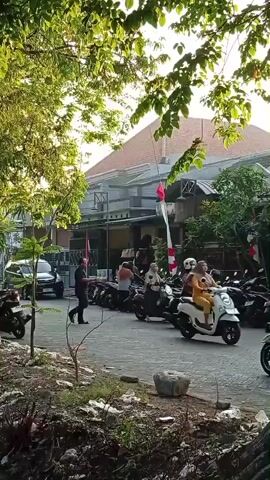 The video shows a street scene with several motorcycles and a few people. The sun is shining brightly, casting long shadows. In the foreground, there is a large tree on the right side of the frame, with its trunk partially obscuring the view of the street. Leaves from the tree hang down from the top of the frame. The street appears to be paved with cobblestones or pavers.

On the street, several motorcycles are parked or being ridden. On the left side, a dark-colored car is partially visible. A person in a dark jacket is walking across the street, away from the camera. Further down the street, several people are riding motorcycles, most of them wearing helmets. One person is riding a scooter with a large bag on the back. Indonesian flags, red and white, are visible on poles attached to buildings in the background. The buildings are low-rise, with tiled roofs. There is some greenery and vegetation along the side of the street and around the buildings. The overall impression is a typical street scene in a residential area, possibly in Southeast Asia, given the presence of the flags and the types of motorcycles.