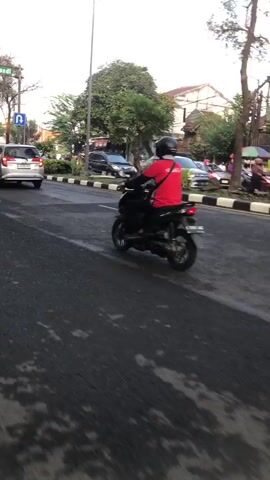 The video captures a street scene in a city. The primary focus is a person riding a black motorcycle, wearing a red shirt and a black helmet. The motorcycle is in motion, moving away from the camera and towards the right side of the frame. The rider appears to be looking forward, possibly navigating the road.

The environment is a paved road with distinct lane markings. The road appears somewhat wet, with reflective patches suggesting recent rain or moisture. On the left side of the road, a silver car is visible, also in motion, moving in the same direction as the motorcycle. Further in the background, other vehicles, including a black car, are present, indicating traffic. Trees and buildings line the street, with a mix of architectural styles visible. The lighting suggests it is either early morning or late afternoon, with a soft, natural light illuminating the scene. A street light pole is also visible. The overall impression is an ordinary urban street with moderate activity.