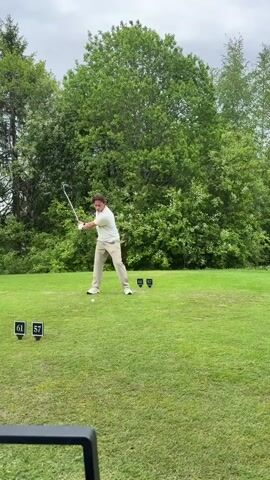 A man in a white shirt and tan pants swings a golf club on a green golf course. The sky is overcast. There are black number signs labeled "61" and "57" in front of him. Green trees and bushes are in the background. He follows through with his swing, looking up after hitting the ball. A black object is partially visible in the bottom left corner of the frame.