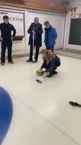 A woman throws a curling stone on an indoor ice rink. She is kneeling, and wearing a dark jacket and dark pants. She lets go of the stone, which is black with a yellow handle. Three people are watching her, two men and one woman. The men are wearing dark jackets. The woman is wearing a blue jacket. A scoreboard is visible in the background. The rink is long and narrow, with white walls. A logo is visible on one wall.