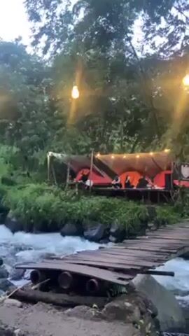 The video shows a scenic outdoor campsite nestled beside a flowing river. In the foreground, a simple, possibly makeshift, wooden bridge spans the river, the water rushing over smooth, dark rocks. On the opposite bank, a raised wooden platform supports several orange tents sheltered under a tarp, lit by string lights. People are visible inside or near the tents, seemingly relaxing. The background is filled with lush green trees and vegetation, partially obscured by the early evening light.