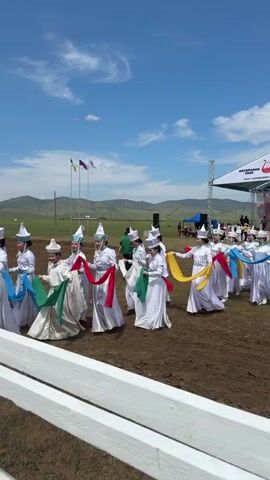 A group of women in traditional white dresses and pointed hats are dancing outdoors. They are holding colorful silk sashes, red, yellow, green, and blue, and moving in a line. The ground is dirt, and in the background, there are hills, a blue sky with white clouds, flags, a stage with a tent, and people watching. The women appear to be performing a cultural dance.