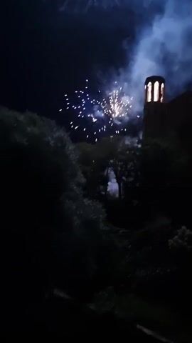 A fireworks display illuminates the night sky behind a building with a tower, surrounded by trees. The fireworks are white and pink, with some sparks and smoke. The building appears to be made of stone and has arched openings in the tower. The trees are dark and silhouetted against the bright sky. The overall scene is dark and atmospheric.
