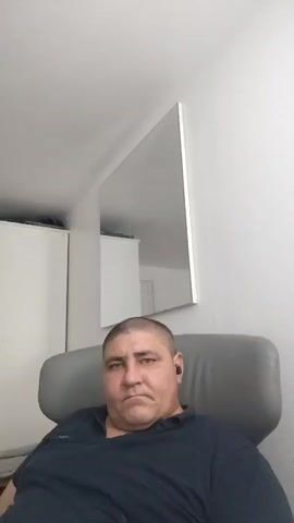A man with a shaved head and a dark shirt sits in a gray chair. He has a small black device in his right ear. Behind him, a large rectangular mirror hangs on a white wall. To the left, part of a white closet or cabinet is visible. The room appears to be lit from above.