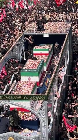 The video shows a large funeral procession with a significant crowd of people. Several coffins are visible on a platform decorated with flowers. The coffins are covered with Iranian flags. The people in the crowd are predominantly wearing black clothing, and many are holding flags. The procession appears to be moving along a street, with buildings visible in the background. The atmosphere seems somber and respectful, with a sense of collective mourning. There are also speakers and audio equipment visible, suggesting that a public address or ceremony is taking place. The overall impression is of a large-scale state funeral or memorial service.