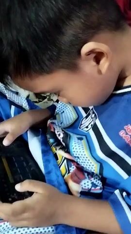 A close-up shot shows a child wearing a blue shirt with a patterned design and dark hair. The child is holding a black tablet in their hands and appears to be playing a game. The tablet screen displays various icons and text, suggesting a game interface. The child's fingers are positioned on the screen, interacting with the game. The background is out of focus, but hints of fabric, possibly bedding or clothing, in blue and white with polka dots are visible. The lighting is dim.