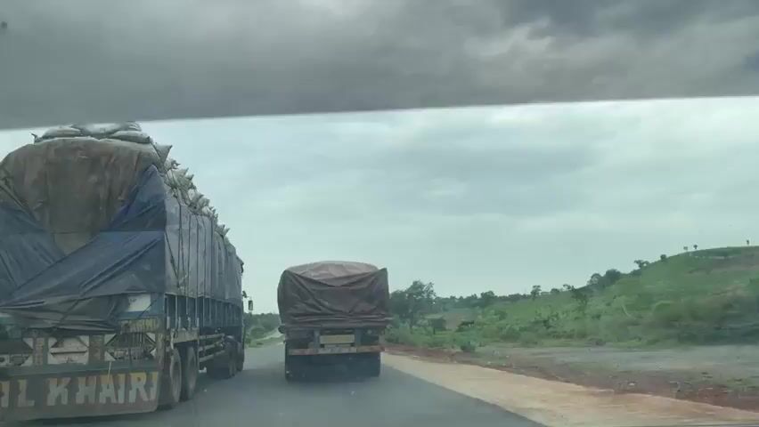 This video depicts a view from inside a vehicle traveling on a road in Kwali, Nigeria. The primary subjects are two large trucks on the road ahead. The truck in the foreground, on the left, is a blue and white semi-trailer truck heavily loaded with what appears to be sacks or bales covered by a blue tarp. The text "AL KHAIRI" is partially visible on the rear of this truck. Further down the road, another truck, a smaller semi-trailer, is visible, also covered with a brown tarp.

The scene is set on a paved road with a dirt shoulder. To the right of the road, a green, hilly landscape with scattered trees and vegetation stretches into the distance. The sky is overcast with dark, heavy clouds, suggesting the possibility of rain or that it has recently rained. The overall mood is subdued due to the weather. The time of day is unclear due to the cloudy conditions, but it appears to be daytime. The activity taking place is transportation, with the trucks likely carrying goods along the highway. The presence of the trucks and the rural landscape are the main visual elements.