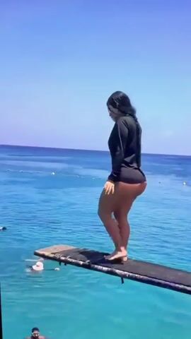 A woman with long dark hair, wearing a black long-sleeved shirt and beige shorts, stands on a wooden diving board over clear blue water. She is facing away from the camera, looking towards the water. The diving board is weathered and appears to be in good condition. There are other people in the water in the background, swimming and enjoying the sunny day. The sky is clear and blue, with a few distant clouds. The scene appears to be a tropical location, possibly a beach or resort.