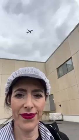 A woman wearing a white cap and red lipstick is taking a selfie video outside, in front of a large, boxy, beige building. An airplane is flying overhead in the cloudy sky. The building has square panels, a few small windows, and a surveillance camera on the corner. There's a "no parking" sign on a low wall in front of the building.
