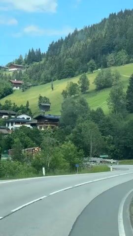 The video shows a view from a vehicle driving on a winding road through a mountainous, forested area. On the left side of the road, there are houses nestled on a grassy hillside dotted with trees. The road itself has white lane markings. There are some road signs visible, including one with the number 21 and a green sign with white text and an arrow pointing right. The sky is blue with some clouds. The overall scene is serene and depicts a scenic drive.