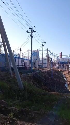 The video pans across an outdoor industrial site on a clear day. The foreground features rough, uneven terrain with dry grass and dirt. In the middle ground, there are several electrical poles with wires strung between them, supporting the power infrastructure for the facility. Behind the poles, a fenced-off area contains multiple industrial buildings and structures. These include what appear to be transformers or electrical distribution units, some with blue exteriors, and a tall, silver smokestack with a ladder extending up its side. The background shows a clear, pale blue sky. There are no people visible in the video, and the only sounds are the ambient noises of the environment. The overall impression is of a functional, albeit somewhat unkempt, industrial power or manufacturing facility.