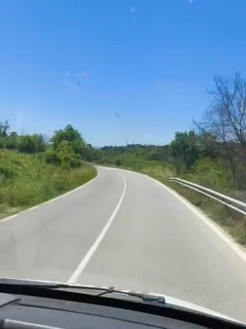 This is a first-person perspective video of a person driving on a paved road on a clear, sunny day. The road is two lanes wide with a white solid line dividing the lanes and white lines on the outer edges. The road curves to the left in the distance. The landscape on either side of the road is lush and green with trees and grass. The sky is a vibrant blue. The windshield of the vehicle appears to be slightly dirty. There are no other vehicles or people visible in the frame. The video lasts for approximately 5 seconds.