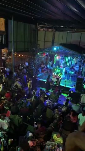 Here is a detailed description of the video:
The video shows a lively, crowded outdoor bar or club at night. Patrons are seated at tables, many with drinks, while others stand and move around. A DJ station is elevated and lit with blue and green lights. People are dancing near the stage. String lights are visible in the background. The space has a high ceiling, and the overall atmosphere is vibrant and social.