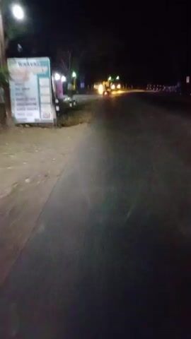 The video is taken at night from a moving vehicle. The road is dark with streetlights illuminating sections of the scene. On the left side of the road, there are a few structures with bright lights, including a sign that says "KARAOKE". There are other vehicles with headlights visible on the road ahead, suggesting it is a moderately trafficked area. The camera angle is low, as if mounted on the dashboard or handlebars. The motion is somewhat shaky, indicating the vehicle is in motion.