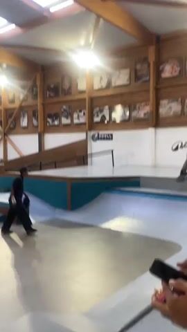 The video shows skateboarders in an indoor skatepark. The park has wooden ramps, platforms, and rails with a light blue trim. There are multiple skaters, one wearing dark clothing, attempting a trick off a ramp but falls. Another skater wearing a light-colored outfit is seen skating around the park. The interior of the skatepark features a wood-beam ceiling with skylights and walls adorned with framed pictures and logos. There is a person holding a phone, possibly recording the skaters. The overall atmosphere appears casual and recreational.
