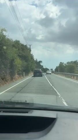 A vehicle travels on a two-lane road. A dark-colored SUV is in front, followed by a white sedan. The road is lined with trees and foliage on the left, and a guardrail on the right. Overhead power lines are visible along the left side of the road. The sky is cloudy with patches of blue. The dashboard of the filming vehicle is partially visible in the foreground.