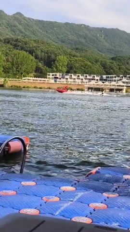 A video begins on a blue floating dock with metal railings and orange grips. The dock is on a body of water. In the distance, there is a red boat or personal watercraft with a person on it, appearing to be waterskiing or jet skiing. The water is rippled. Behind the water, there are green, tree-covered mountains under a bright, partly cloudy sky. In the mid-ground, there is a shoreline with buildings and more trees. A yellow sign is partially visible on the dock in the foreground.