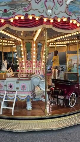 A carousel with ornate decorations and lights is in motion. A young boy in a yellow shirt stands on the carousel. Two children, a girl and a boy, both wearing red shirts, are on the sidewalk next to the carousel. The girl appears to be holding a pole connected to the carousel, and the boy is standing beside her. Other people and vehicles are visible in the background, suggesting an urban setting.
