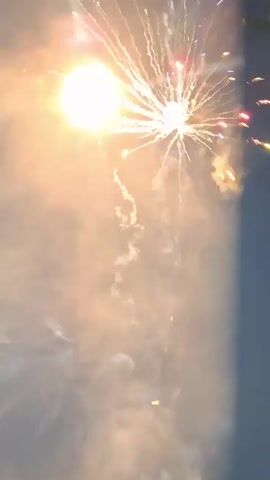 This video captures a fireworks display at night. The fireworks are primarily golden and orange, bursting in the dark sky, with some smoke visible. The display appears to be in stages, with various bursts and falling embers captured at different times. The lower portion of the frame is darker, with some foliage or trees visible on the right side in one frame.