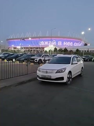 The video shows a parking lot in front of a stadium at dusk. A white car is parked in the foreground. In the background, the stadium is lit up with colorful lights, displaying text that appears to read "Сочи" or something similar in Russian. Several other cars are parked in the lot, and a fence separates the parking area from the stadium. A sign on the fence reads "ВЫЕЗД ЗАПРЕЩЕН," meaning "EXIT PROHIBITED" in Russian. The sky is a dusky blue, indicating it is either early evening or late afternoon.