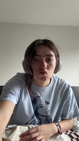 A woman in her late teens or early twenties is seated on a bed with a brown and white checkered pattern. She wears a light blue t-shirt with a print on it, and grey headphones. She is opening a beige cotton drawstring bag. The walls of the room are plain white.