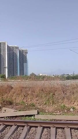The video shows a view from a moving train, with a focus on the landscape outside. In the foreground, the train tracks are visible, along with gravel and dry vegetation. Beyond the tracks, there's a field of brown grass with scattered green plants. In the background, there are multiple high-rise buildings under construction. They are gray and incomplete, with some covered in blue netting. The sky is clear and blue. The camera moves along with the train, giving a sense of travel through the landscape.
