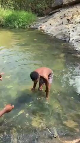 The video shows a young man standing in a clear, shallow river. He is dark-skinned, shirtless, and wearing patterned shorts. He is smiling. Another person, only partially visible, splashes water at him. The river is surrounded by rocks and greenery. The water appears clean, with some rocks visible beneath the surface.