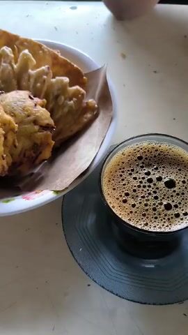 The video shows a white plate with a pile of fried food resting on a brown paper liner.  Next to the plate is a clear glass of dark coffee with bubbles. The coffee glass rests on a gray saucer. The scene is shot from a slightly elevated angle, showing the food and drink on a light-colored surface. An arm is briefly visible in the upper right corner.