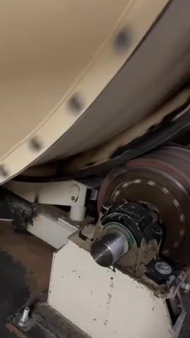 The video shows a close-up view of a machine that appears to be processing a large roll of material. A beige-colored material, possibly paper or fabric, is being fed through the machine. Dark circular marks are visible on the surface of the material. The material is guided by rollers, one of which is visible in the foreground. This roller is attached to a bearing housing which is covered in a light-colored dust or debris, possibly wood or paper dust. The roller itself is rotating, indicated by the slight blurring of its surface. The machine appears to be industrial in nature, and the environment seems to be a workshop or factory. The lighting is consistent with indoor industrial settings.