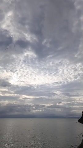 The video shows a wide expanse of water, likely a large lake or ocean, under a cloudy sky. The clouds are varied, with some appearing dark and heavy, while others are lighter and more scattered, allowing patches of sunlight to break through. The water's surface is relatively calm, with gentle ripples. On the right side of the frame, a small portion of land is visible, with what appears to be a treeline. The overall lighting suggests it might be early morning or late afternoon, as the sun is not directly visible. There are no people or discernible objects in the water, and the scene is tranquil.