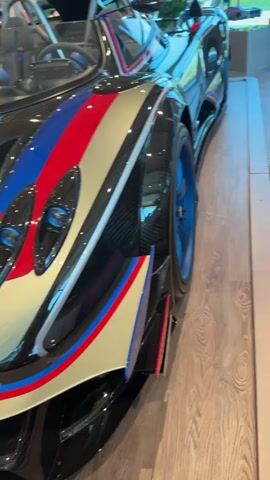 The video pans across the side and interior of a silver Pagani Huayra Roadster BC, a high-performance sports car. The car is parked on a polished wooden floor in what appears to be a showroom or dealership. The exterior features a silver finish with red, white, and blue racing stripes on the side and a prominent rear wing. The wheel rims are blue and black. Inside, the car has a luxurious interior with brown leather and carbon fiber seats, a sleek steering wheel, and a detailed dashboard. Two people, a man in a blue t-shirt and a woman with short red hair, are visible in the background, looking at the car. The environment seems to be a well-lit indoor space with display cases in the background.