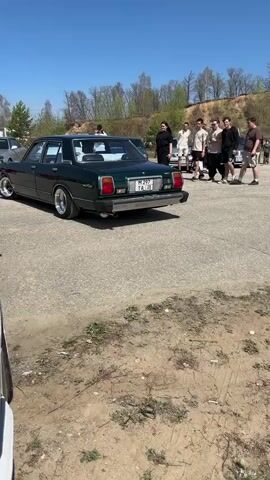 The video shows a dark green, vintage Toyota Mark II sedan parked on an asphalt and dirt lot under a bright blue, cloudless sky. A group of approximately twelve people are standing on a slight incline near the car, conversing casually. A white Toyota sedan is parked a distance away from the green car. The license plate of the green car, which is mounted on the trunk, displays "M 297 TA 28". The car is parked near the edge of the asphalt, with dirt and small patches of sparse greenery visible closer to the camera. Trees and other vegetation populate the background, along with a dirt hill.