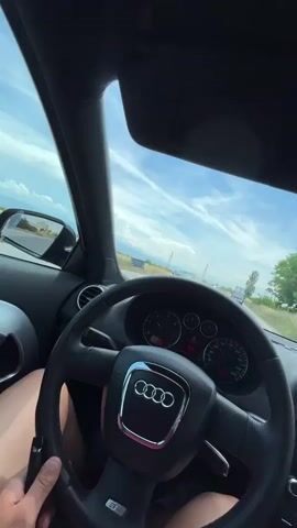 The video provides a first-person perspective from inside a car. The driver's hands are visible on the steering wheel, which has an Audi logo. The car's dashboard, including the speedometer and tachometer, is in view. The road ahead is visible through the windshield, with other cars in the distance. The sky is blue with white clouds. The scene appears to be a daytime drive on a rural or suburban road, with vegetation and utility poles lining the sides. The car's audio system display shows the word "ENERGY."