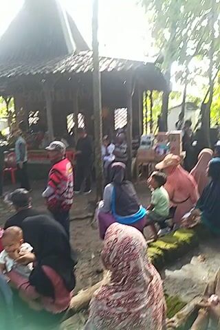 Here is a concise description of the video:

The video shows a gathering of people in an outdoor setting, possibly a village or rural area. Many are standing, sitting, or kneeling on the ground. Some are under the cover of a simple wooden structure. Some individuals are wearing traditional or casual clothing, and a woman is holding a baby.  The video is shot from a high angle, and the composition is focused on the group of people. The environment includes trees and other greenery. There is music playing in the background.