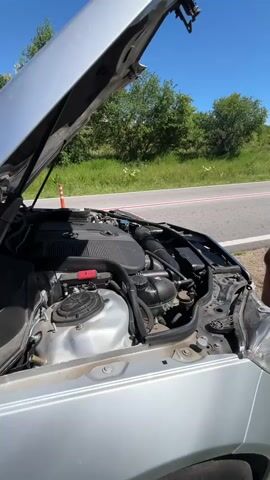 The video shows a silver Mercedes-Benz with its hood open on the side of a road. The car's engine is visible, and a person is standing next to the car, possibly examining the engine. The road has a red and white stripe near the edge, and there is a red cone nearby. The background features trees and greenery under a clear blue sky.
