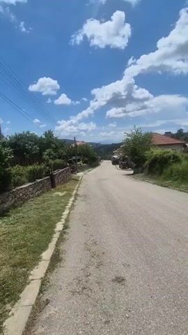 The video shows a street in a village on a sunny day. The street is paved and there are houses on both sides. There are trees and grass on the sides of the street. The sky is blue with white clouds. There are two cars parked on the side of the road.