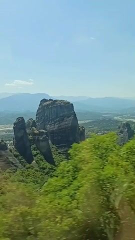The video captures a scenic view of large, towering rock formations covered in lush green vegetation. The sky is clear and blue, with distant mountains visible. The camera pans across the landscape, highlighting the dramatic geological features. The speaker mentions that the rock formations resemble candles and implies that someone may have died there, though this is not visually depicted in the video.