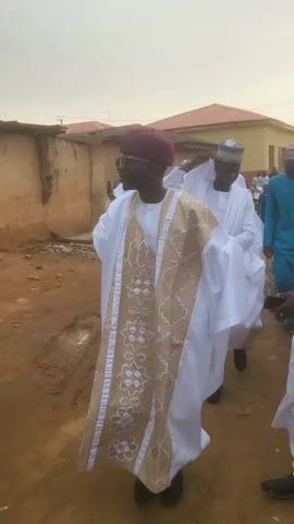 A group of men is seen walking in a procession outdoors in Jogana, Nigeria. The men are predominantly dressed in traditional white robes, resembling kaftans or agbadas, many featuring intricate gold or patterned embroidery. They wear various traditional caps, such as filla or kufi.

In the foreground, a man with a white robe adorned with a prominent gold and orange patterned design on the front wears a distinctive red and white striped cap. Beside him, another man sports a maroon cap and sunglasses, with a richly gold-patterned sleeveless garment over his white attire. Several men also wear sunglasses, and one man holds a mobile phone.

The setting appears to be a street or pathway, with simple buildings, including one with a red roof, and utility poles or power lines visible in the background. The sky is bright, suggesting daytime, possibly late afternoon or early evening, with diffused light. The men appear composed as they walk, indicating an orderly movement, potentially part of a cultural, religious, or celebratory event. No discernible text is visible in the frame.