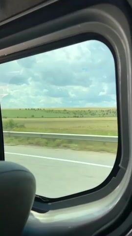 The video is a first-person view from inside a vehicle looking out a window. The window is oval-shaped and shows a landscape of rolling hills and fields. The sky is cloudy and blue. A road is visible in the foreground, and trees are scattered across the fields. The vehicle appears to be moving, as the landscape outside is slightly blurred.