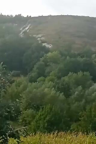 The video shows a rural landscape. In the foreground, there are plants that appear to be crops, possibly corn, with some green foliage around them. In the middle ground, there is a dense area of green trees and bushes. In the background, there is a hillside covered with greenery and some lighter colored patches that might be rocks or bare earth. The sky above is overcast with some clouds. The camera appears to be moving, panning across the scene. There are no people or animals visible. The overall impression is of a natural, possibly agricultural, environment.