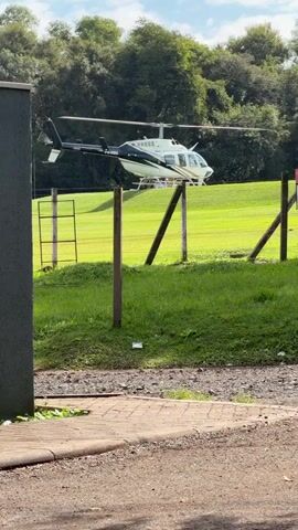 A white and black helicopter is the primary subject of this short video. The helicopter is stationary, hovering slightly above a grassy field. It appears to be either landing or taking off. The setting is outdoors with a clear sky and lush green trees in the background. A fence made of wooden posts and wire runs across the foreground, partially obscuring the view of the helicopter. A small section of a brick pathway is visible in the bottom left corner, suggesting a constructed area nearby. The overall scene appears to be a rural or semi-rural location, possibly a private helipad or a field adjacent to a property. The time of day seems to be daytime, with bright lighting indicating sunshine. No people are visible in the frame. The registration number "PR-HBU" is visible on the side of the helicopter.