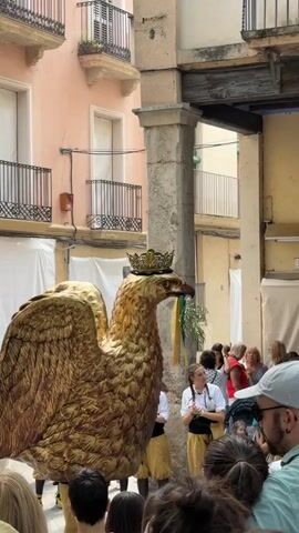 A giant golden eagle costume with a crown on its head is being paraded through a narrow street. The eagle has its wings spread and is holding a branch with green leaves and colorful ribbons in its beak. A young person dressed in a white shirt and gold pants is walking behind the eagle, holding onto part of the costume. Several other people, some holding babies, are watching the procession from the sides of the street. The buildings lining the street have balconies with decorative railings. The sky is not visible. The scene appears to be part of a festival or parade.