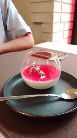 The video shows a person seated at a table in what appears to be a restaurant or cafe. On the table, there's a black plate with a clear, round glass bowl containing a dessert with a white base and a bright pink topping, decorated with small flowers. A silver spoon rests on the plate. Next to the plate is a cell phone. The camera then shifts focus to a gold-colored candle holder and a window with a view of evergreen trees outside. The window is framed with dark wood, and there's a brick wall with red lighting visible on the left side of the frame.