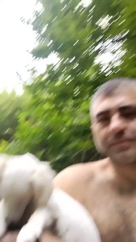 A man is holding a small white dog. The dog is looking down and appears to be nibbling on the man's arm. The man is smiling at the camera. In the background, trees are visible, and there is a waterfall and a stream. The man appears to be shirtless.