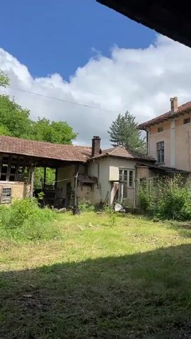 The video presents a serene, daytime scene in Byala Reka, Bulgaria, capturing an old, rustic house under a partly cloudy sky. The initial wide shot at 0:00-0:01 displays the house with a traditional tiled roof and a rough, stucco-like exterior. A brick chimney stands prominently on the roof. The house is surrounded by a green, slightly overgrown grassy yard. To the left, lush green deciduous trees are visible, while a tall pine tree stands to the right, partially obscuring another building. Overhead, power lines cut across the sky.

As the video progresses from 0:01-0:03, the camera slowly zooms in, gradually narrowing the focus to the house's roof and chimney. The details of the aged, terracotta-colored roof tiles become clearer, showcasing their weathered texture. The brickwork of the chimney is distinct, appearing robust against the backdrop of the sky. The final frames focus tightly on the chimney and the surrounding roofline, with the mix of bright blue sky and fluffy white clouds dominating the background, framed by the top of the pine tree. The overall impression is one of rural tranquility and a glimpse into traditional architecture. No visible text is present in the video.