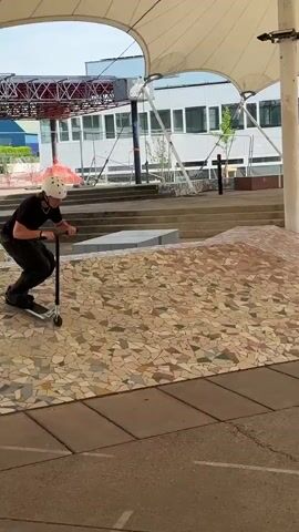 A young man riding a scooter attempts a trick on a staircase but falls. The man is wearing a black shirt, black pants, and a white helmet. The scooter is silver and black. He approaches a short set of stairs, jumps off the first one, but loses his balance and falls to the ground on the next level. The scooter breaks in half. The background includes a building with large windows, some construction, and a tiled surface. The video is shot outdoors during the daytime.