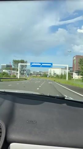 The video shows a driver's perspective from inside a car approaching a road intersection. The car is stopped, likely at a red light.  The camera faces a road intersection with clear signage indicating directions to "Leiden-Centrum" and "Bio Science Park-Oost I and II". The day is bright with a partly cloudy sky.  Modern buildings are visible in the background, typical of a business park or city outskirts. The vehicle's dashboard is visible in the foreground, displaying an "SRS AIRBAG" indicator. The traffic light ahead is green.