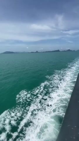 The video shows a view from the side of a moving boat, looking out at the ocean. The water is a vibrant turquoise color and is choppy, with white foam trailing behind the boat. In the distance, a faint outline of islands or a coastline can be seen against a cloudy blue sky. The edge of the boat is visible on the right side of the frame. The lighting suggests it is daytime, likely under a partly cloudy sky. The camera appears to be held steady, capturing the motion of the water.